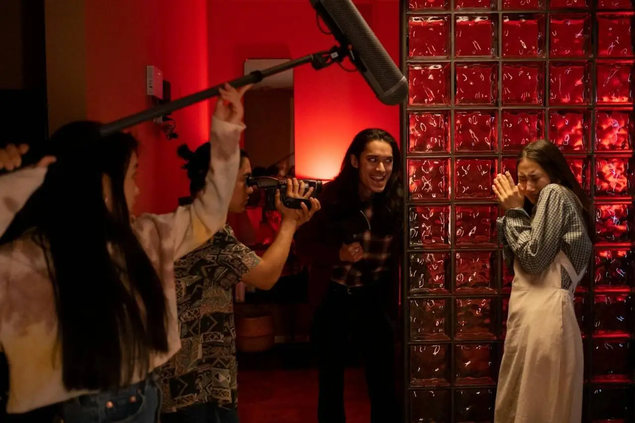 A group of teenagers filming a playful horror scene indoors with a boom mic and camera.